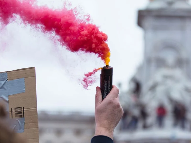 Una mano sostiene una bengala de humo rosa durante una manifestación, con un monumento al fondo.
